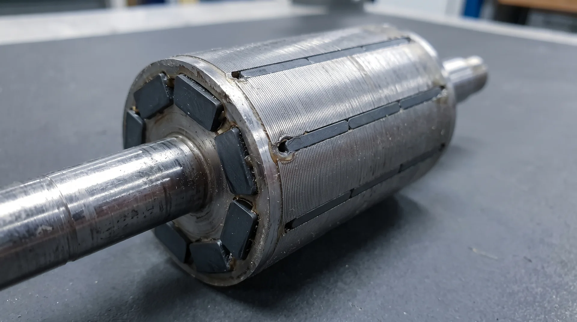 A cylindrical laminated motor rotor lying on a bench, with dark magnet blocks set into slots around its shaft.