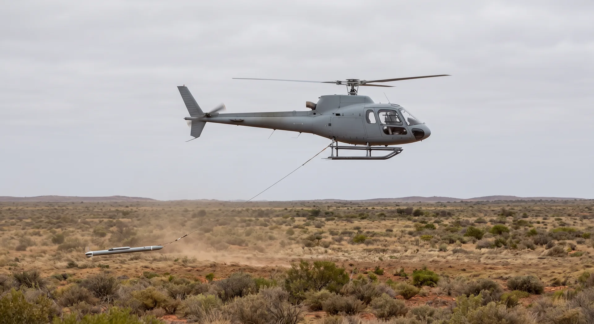 A helicopter flying low over open scrubland towing a long cylindrical sensor on a trailing cable that rides just above the ground.