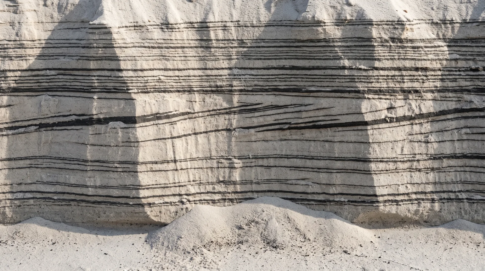 A cut face through beach sand banded with many thin dark layers, some continuous and some lensing out, truncating against each other partway up.