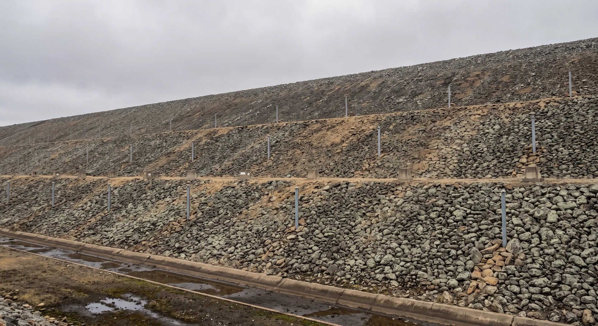 The rock-armoured downstream face of a dam embankment rising in two benched stages, slim standpipes set into it at intervals and a drainage channel at the toe.