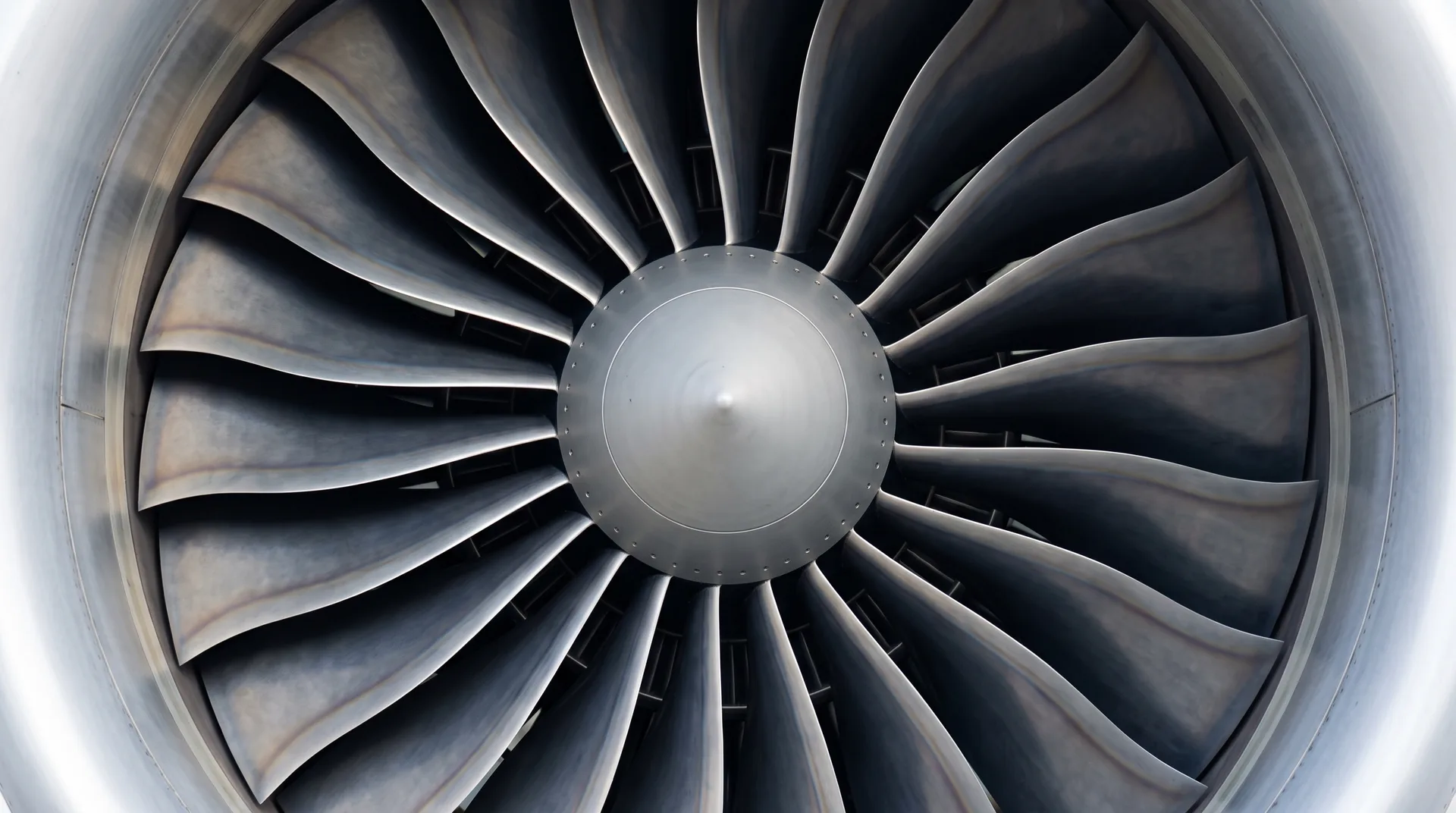 The front fan of a jet engine seen head-on, its broad curved blades radiating from a central spinner inside a bright nacelle.