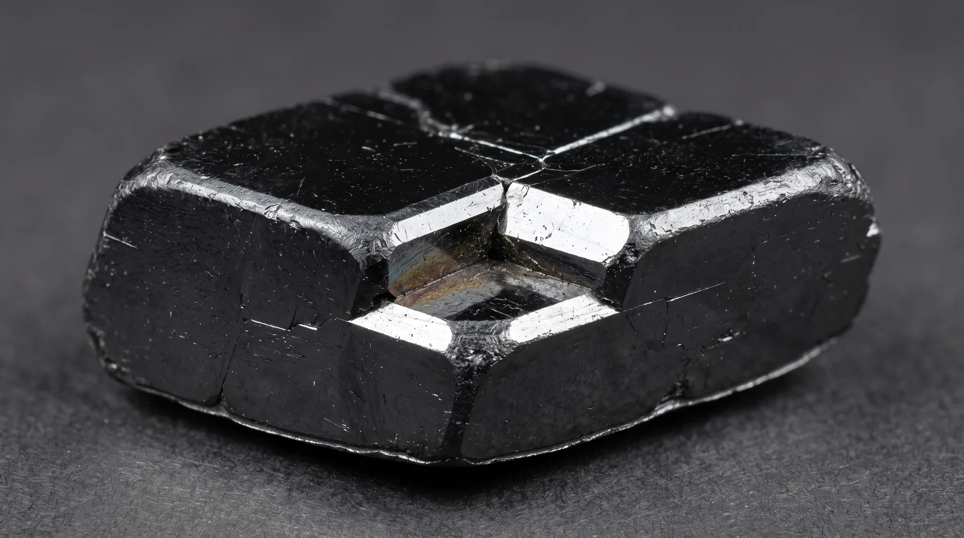 A blocky black crystal with bright angular cleavage faces and a metallic sheen, on a dark grey ground.