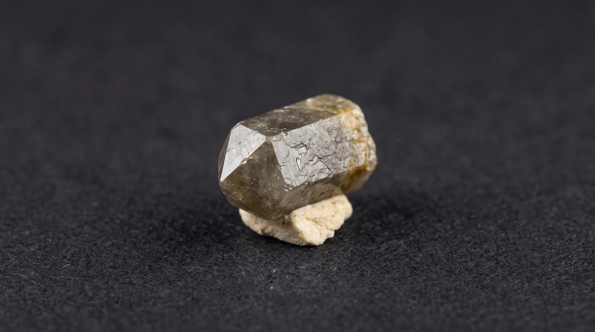 A small pale grey-brown crystal with a short prismatic form and angled end faces, perched on a chip of light matrix.