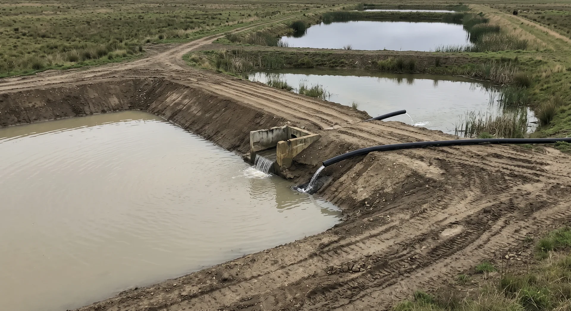 Three settling ponds stepping away in line between earth bunds, the nearest water turbid and the furthest still and reflecting the sky.
